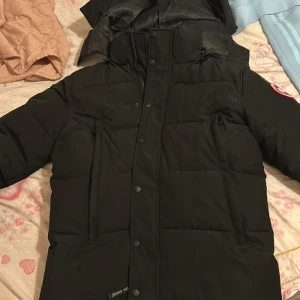 Svart dunjacka från Canada Goose M - Säljer en svart dunjacka från Canada Goose i storlek M. Jackan har huva, klassisk logga på ärmen och stängs med både dragkedja och knappar. Perfekt för kalla vinterdagar och riktigt snygg streetstil.