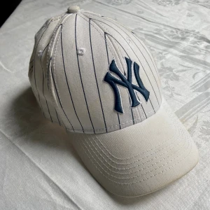Vit New York Yankees keps - Snygg vit keps från New York Yankees med tunna svarta ränder och broderad NY-logga framtill. Justerbar snapback baktill och texten 'New York' broderad ovanför spännet. Perfekt accessoar för en sportig look.