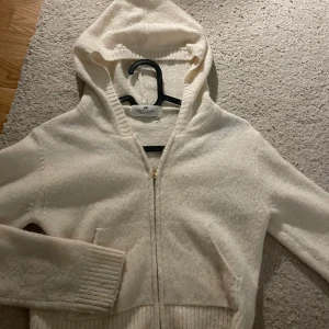 Vit hoodie med dragkedja från H&M - Mysig vit hoodie från H&M i storlek 134/140. Tröjan har huva, ribbade muddar och praktiska fickor framtill. Materialet är stickat och mjukt, perfekt för kyliga dagar. Stängs med en snygg guldfärgad dragkedja framtill.