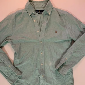 Grön skjorta från Ralph Lauren S - Snygg grön skjorta från Ralph Lauren i slim fit med klassisk krage och broderad logga på bröstet. Skjortan har långa ärmar, vita knappar och är gjord i mjuk bomull. Perfekt för dig som gillar stilrena och tidlösa plagg. Tappat lite färg på magen zooma in för att se