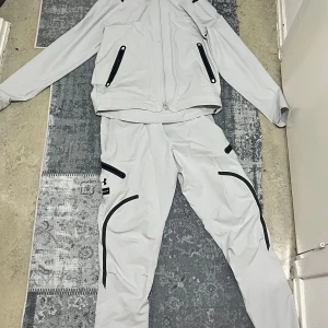 Under Armour ljusgrå träningskostym - Säljer en ljusgrå träningskostym från Under Armour med svarta detaljer och dragkedjor. Setet består av en jacka med hel dragkedja och matchande byxor med flera fickor och logga på benet. Perfekt för dig som gillar sportig och modern stil.