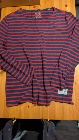 Randig långärmad tröja Cottonfield XL - Snygg långärmad tröja från Cottonfield i storlek XL. Marinblå och röd med horisontella ränder, rund hals och en cool patch med siffran 86 nedtill. Tillverkad i mjuk bomull, perfekt för dig som gillar avslappnad stil. Obs: ett litet riv längst ned på vänstra armen(vid handleden), enkelt dolt med ett litet vik upp armen. 