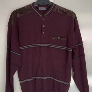 Vinröd stickad tröja Martinelli medium - Vinröd stickad tröja från Martinelli med bruna mockadetaljer på axlar och bröstficka. Tröjan har knappar vid halsen, ribbade muddar och diskreta ränder i beige och blått. Perfekt för dig som gillar retrovibbar och snygga detaljer.