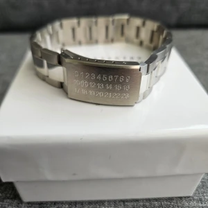 Maison Margiela silverarmband - Coolt armband från Maison Margiela i silverfärgat rostfritt stål. Länkarna är breda och matta med en unik graverad platta med siffror, där 11 är markerad. Perfekt accessoar för dig som gillar stilrena och moderna detaljer.