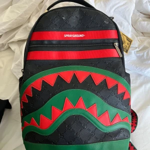 Sprayground limited edition ryggsäck - Limited edition ryggsäck från Sprayground med svart bas, röda och gröna detaljer samt ikoniskt hajtandsmönster. Väskan har präglat mönster, flera fack med dragkedja och justerbara axelremmar. Perfekt för dig som vill sticka ut med en unik och färgstark accessoar.