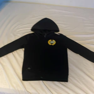 Svart Hammarby hoodie med dragkedja - Svart hoodie från Hammarby med klubbens emblem i gult och grönt på bröstet. Tröjan har huva, hel dragkedja framtill och långa ärmar. Perfekt för dig som vill visa ditt stöd för laget och hålla stilen.