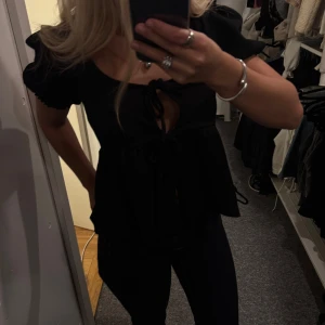 Svart blus med puffärm och knyt - Supersöt svart blus med korta puffärmar och två knyt framtill som ger en snygg detalj. Blusen har en lätt utsvängd passform och djup urringning.