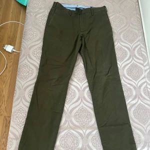 Chinos från Polo Ralph Lauren & H&M - Två par chinos, ett i olivgrönt från Polo Ralph Lauren med klassisk passform och bälteshällor, samt ett par i beige från H&M. Båda har raka ben och är tillverkade i bomull, perfekta för en clean och stilren look.