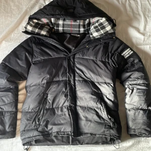 Svart Burberry pufferjacka M,  - Svart pufferjacka från Burberry med stor huva och rutig insida i svart, vit och röd. Jackan har två dragkedjefickor, tydlig Burberry-logga på ärmen och quiltad design. Perfekt för kalla dagar och har en riktigt snygg streetstil. PRIS KAN DISKUTERAS 