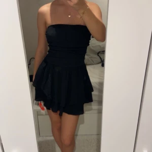 Svart axelbandslös kortklänning Cider - Supersnygg svart axelbandslös kortklänning från Cider i storlek XS. Klänningen har volangig kjol och sitter tajt upptill. Tillverkad i 100% polyester och perfekt för dig som vill ha en stilren och trendig look.