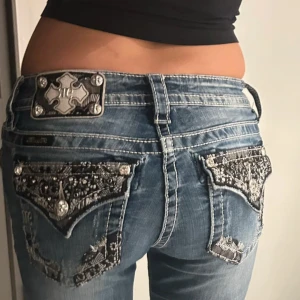 Miss Me jeans💕 - Snygga blå Miss Me jeans med låg midja, dekorerade bakfickor med svart spets, nitar och broderade detaljer. Jeansen har slitningar och kontrastsömmar, samt en unik metallplatta bak i midjan. Modellen har bootcut och är low waist och coola detaljer som verkligen sticker ut.
