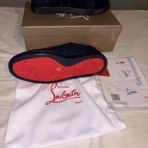 Christian Louboutin marinblå sneakers - Exklusiva marinblå sneakers från Christian Louboutin i mocka med ikonisk röd sula. Skorna har rund tå, snörning och diskret logga på sidan. Insidan är i ljusbeige läder och sulan har Louboutin-signatur. Perfekt för dig som vill sticka ut med stil.