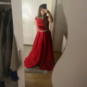 Röd långklänning med glittrigt bälte - Säljer en röd långklänning med offshoulder-modell och glittrigt bälte i midjan. Klänningen har en elegant, vid kjol och markerad midja. Perfekt för bal eller festliga tillfällen. Materialet känns slätt och faller snyggt.