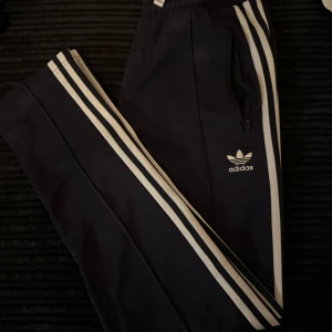 Adidas mjukisbyxor  - Marinblå mjukisbyxor från Adidas med klassiska vita ränder längs benen och broderad logga på låret. Byxorna har resår i midjan och dragkedjeförsedda fickor. Perfekta för en sportig och avslappnad stil.