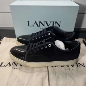 Marin blåa sneakers från Lanvin i mocka - Snygga svarta sneakers från Lanvin med ovandel i mocka och lackad tå. Skorna har vita platta sulor och svarta snören. Klassisk och stilren design med rund tå och metallfärgade öljetter. Perfekta för dig som gillar exklusiva och trendiga sneakers. Det är storlek 46 om jag inte har fel
