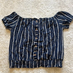 Randig offshoulder topp från H&M - Mörkblå offshoulder topp från H&M med vita ränder och knappar framtill. Toppen har korta ärmar och resår vid axlar och nederkant för en snygg passform. Perfekt för en avslappnad och trendig look. Kontakta för fler bilder🩷