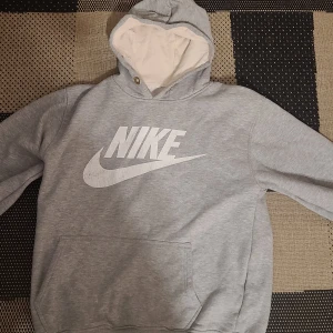 Grå Nike hoodie med stor logga - Grå hoodie från Nike med stor vit logga framtill. Klassisk känguruficka och huva med vit insida. Perfekt för chill dagar eller när du vill ha en sportig look. Mjuk och skön med lång ärm och relaxed fit.