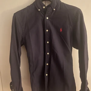 Mörkblå skjorta från Ralph Lauren - Snygg mörkblå skjorta från Ralph Lauren i slim fit-modell. Skjortan har klassisk button down-krage, vita knappar och den ikoniska röda broderade loggan på bröstet. Byten mot liknande skjortor i strl M är också intressant. 
