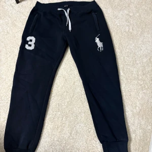 Mörkblå mjukisbyxor från Ralph Lauren - Mörkblå mjukisbyxor från Ralph Lauren med vit snörning i midjan, broderad stor logga och siffran 3 på ena benet. Byxorna har ribbade muddar vid bensluten och två fickor framtill. Perfekta för en chill och sportig look.