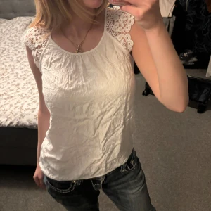 Vit blus med spets från Atmosphere - Supersöt vit blus från Atmosphere med broderade spetsdetaljer på axlarna och ärmlös design. Blusen har en lätt struktur i tyget och knäppning bak i nacken. Perfekt för dig som gillar romantisk och feminin stil. Den e lite skrynklig på bilden, så den behöver strykas men annars är den så himla fin så va lite osäker på om jag ville sälja. Den har jättefint mönster på tyget, vilket jag älskar!