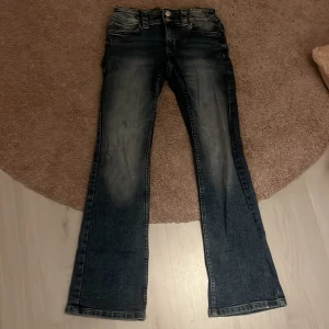 Bootcut jeans från H&M, blå denim - Bootcut jeans från H&M i blå tvättad denim med snygga slitningar och dubbla knappar i midjan. Klassisk femficksmodell med ficklock och knappar bak. Jeansen har en lätt utsvängd passform och coola detaljer som ger en trendig look.
