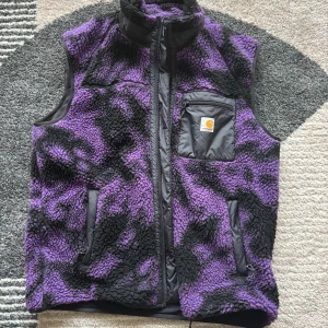 Lila och svart fleeceväst från Carhartt - Säljer en fet fleeceväst från Carhartt i lila och svart camouflage-mönster. Västen har hög krage, dragkedja framtill och tre fickor med zip, varav en bröstficka med Carhartt-logga. Perfekt för lager-på-lager och chill stil.