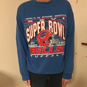 Blå Buffalo Bills Super Bowl tröja - Retro blå crewneck från Trench med stort Buffalo Bills Super Bowl-tryck i rött, vitt och blått framtill. Skön passform med långa ärmar och ribbade muddar. Materialet är en mix av bomull och polyester, perfekt för dig som gillar amerikansk sportstil.