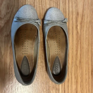 Grå ballerinaskor från Ecco i skinn - Snygga grå ballerinaskor från Ecco i mjukt skinn med rund tå och dekorativ rosett framtill. Ovandelen har ett diskret mönster och innersulan är i brunt läder för extra komfort. Perfekta för dig som gillar stilrena och bekväma skor.