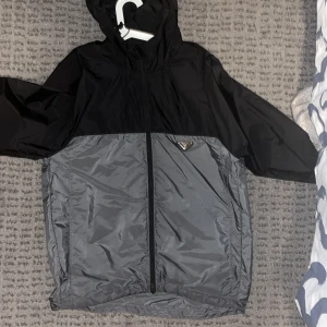 Svart och grå vindjacka från Prada - Snygg vindjacka från Prada i svart och grått med huva och hel dragkedja framtill. Jackan har en clean tvåfärgad design och ett diskret Prada-emblem på bröstet. Perfekt för dig som gillar stilren streetwear och vill ha något som funkar när det blåser. Autentisk!!!!
