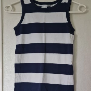 Randigt linne från My Wear Young - Snyggt randigt linne från My Wear Young med breda marinblå och vita ränder.. Tillverkad i mjuk bomull som känns skön mot huden. Rund halsringning och klassisk design som ger en fräsch look. Liten fläck på linnets fåll (se bild). Jag har inte försökt att få bort den så det är mycket möjligt att den försvinner om man behandlar den.