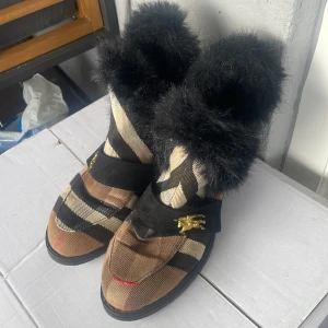 Lyxiga Rutig Burberry boots med svart fuskpäls - Unika Burberry liknade boots med rutigt mönster i beige, brunt, svart och rött. Skorna har svart fuskpäls upptill och svarta detaljer i mocka med guldfärgade emblem. Platt sula och rund tå ger en cool och bekväm look. Perfekta för dig som vill sticka ut. 