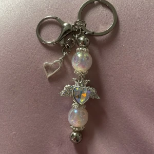 Nyckelring med pärlor och hjärta - Superfin nyckelring i silverfärg med skimrande pärlor, glittrigt hjärta och små vingar. Hängsmycket har en söt hjärtberlock och gnistrar i regnbågens färger. Perfekt accessoar till väskan eller nycklarna för en extra touch av cuteness.