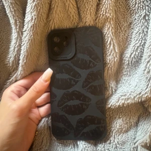 iPhone 12 - Snygg iPhone 12 i svart med dekorativt läpp-mönstrat skal. Telefonen ser ut att vara i gott skick utan synliga repor eller skador på baksidan. Perfekt för dig som vill ha en stilren och trendig mobil. Skalet skyddar effektivt mot stötar och repor.