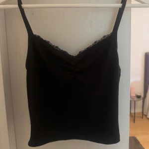 Svart ribbad linnetopp med spets - Snygg svart linnetopp med smala axelband och v-ringning dekorerad med spets. Toppen har ribbad struktur och en lätt rynkning framtill som ger en fin detalj. Perfekt att styla med jeans eller kjol för en trendig look.