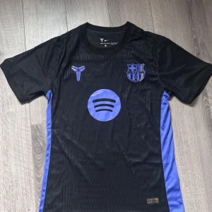 FC Barcelona Lamine Yamal Player Version - Snygg svart och blå FC Barcelona fotbollströja från Nike med Lamine Yamal och nummer 10 på ryggen. Tröjan har korta ärmar, klubbmärke och sponsortryck på bröstet samt blå sidopaneler. Tillverkad i lätt och ventilerande material, perfekt för match eller träning.