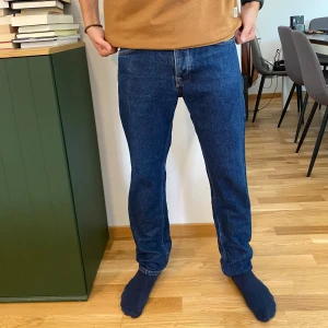 Mörkblå jeans från Nudie Jeans - Snygga mörkblå jeans från Nudie Jeans med orange sömmar på bakfickorna. Jeansen har rak passform, knappgylf och är tillverkade i robust denim av bomull. Sparsamt använda, storlek W31 L30