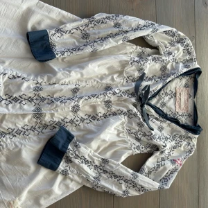 Vit och blå mönstrad klänning Odd Molly - Supersöt vit kortklänning från Odd Molly med blå broderat mönster och blå detaljer vid ärmslut och hals. Klänningen har lång ärm, knytband vid halsen och är gjord i mjuk bomull. Perfekt för dig som gillar boho-stil och snygga detaljer.