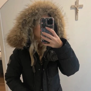 Hollies jacka livigno  - Snygg svart dunjacka med stor fluffig fuskpäls runt luvan. Jackan har dragkedja framtill, flera fickor och markerad midja för en schysst siluett. Perfekt för kalla vinterdagar när du vill hålla dig varm. Nypris ca 3000-4000