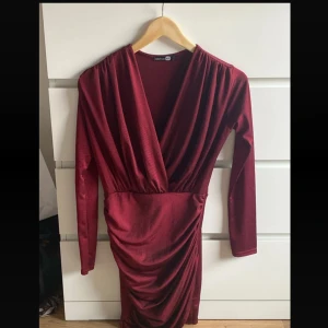 Vinröd omlottklänning från Boohoo - Snygg vinröd kortklänning från Boohoo med omlottmodell och v-ringning. Klänningen har långa ärmar och draperad kjol som ger en cool och trendig look. Perfekt för dig som vill sticka ut med en elegant men edgy stil.