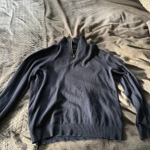 Mörkblå halvzip stickad tröja - Mörkblå stickad tröja med halv dragkedja i halsen. Tröjan har ribbade muddar vid ärmslut och nederkant samt hög krage. Perfekt för kyliga dagar och enkel att matcha med jeans eller chinos.
