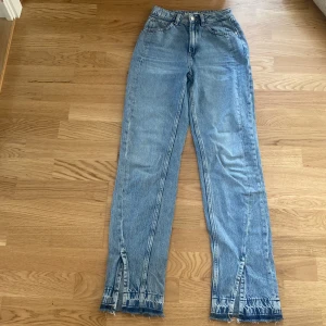 Blå jeans med slits och rå kant - Säljer ett par ljusblå jeans med hög midja och raka ben. Jeansen har coola slitsar och råa, fransiga kanter längst ner på benen för en trendig look. Klassisk femficksmodell och tillverkade i jeansmaterial. Perfekta för dig som gillar en avslappnad men edgy stil.