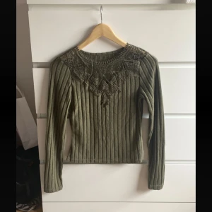 Olivgrön ribbad tröja med spets - Supersnygg olivgrön ribbad tröja med lång ärm och detaljerad spets runt halsen. Tröjan har en figurnära passform och är perfekt för dig som gillar unika detaljer. Materialet känns mjukt och stretchigt.