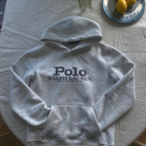Grå hoodie från Polo Ralph Lauren - M/S     Snygg ljusgrå hoodie från Polo Ralph Lauren med stor logga framtill. Klassisk känguruficka, huva och mjukt material som känns skönt mot huden. Perfekt för dig som gillar stilren och avslappnad look.