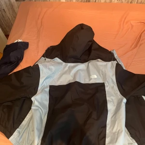 Svart och ljusblå vindjacka The North Face - Vänstra fick dragkedjan är sönder