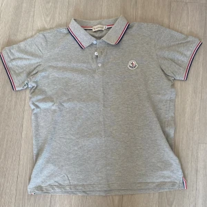 Grå Moncler pikétröja! - Snygg grå pikétröja från Moncler med klassisk krage och knäppning framtill. Tröjan har Moncler-logga på bröstet och detaljer i rött, vitt och blått på krage och ärmslut. Perfekt för en clean look.