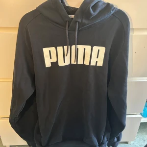 Svart hoodie från Puma med logga - Svart hoodie från Puma med stor vit logga framtill. Tröjan har huva med snörning och är tillverkad i mjukt material med meshfoder i huvan. Perfekt för en sportig och avslappnad stil.