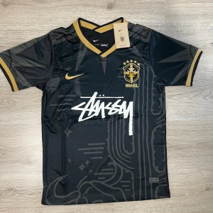 Svart Brasil x Stüssy Nike fotbollströja M - Snygg svart Brasil fotbollströja från Nike i samarbete med Stüssy. Tröjan har guldiga detaljer på krage, ärmslut och loggor, samt unikt grafiskt mönster och Stüssy-tryck på bröstet. Tillverkad i lätt och ventilerande polyester med korta ärmar.