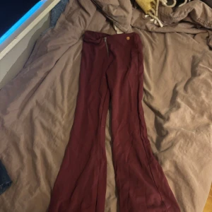 Vinröda bootcut byxor med dragkedja - Snygga vinröda byxor med bootcut-snitt och dragkedja framtill. Byxorna har en hög midja och är tillverkade i ett mjukt tyg som ger en skön känsla. Perfekta för dig som vill sticka ut med en cool färg och trendig passform.