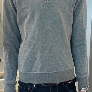 Grå half zip tröja från GANT - Snygg grå tröja från GANT med half zip och hög krage. Tröjan har lång ärm, ribbade muddar och liten broderad GANT-logga på bröstet. Perfekt för dig som gillar klassisk och stilren look. Fläck på arm och på magen (Se bild)