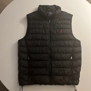 Svart dunväst från Polo Ralph Lauren storlek M - Snygg svart dunväst från Polo Ralph Lauren med quiltad design och dragkedja framtill. Västen har två fickor med dragkedja och klassisk röd logga broderad på bröstet. Perfekt för lager-på-lager och stilren streetlook.